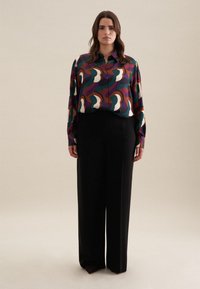 Camicia a maniche lunghe multicolore con motivi astratti in viola, verde e crema, abbinata a pantaloni neri a gamba larga con vita alta.