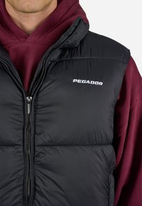 Crna puffer prsluk s visokim ovratnikom, prednjim patentnim zatvaračem i istaknutim bijelim logotipom "PEGADOR". Nosi se preko bordo dukserice s kapuljačom.