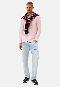Camisa de botones a rayas rosa claro, jeans azules desgastados y un suéter a rayas azul marino caído sobre los hombros. Zapatillas negras completan el atuendo.