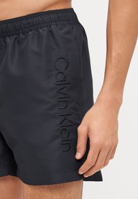 Shorts de bain noirs en matériau léger, avec une taille élastique et un logo "Calvin Klein" brodé sur le côté.