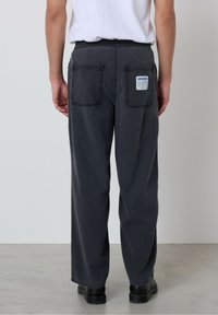 Oseba stoji, oblečena v ohlapne temno sive hlače z elastiko v pasu in zadnjimi žepi, belo majico in črne čevlje na sivi tleh.