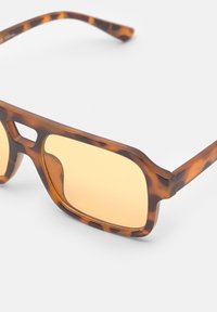 Lunettes de soleil écaille de tortue avec une monture géométrique, des verres dorés et des branches texturées. Matériau plastique léger avec une finition mate.