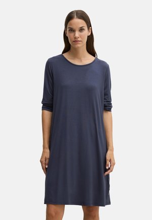 Vrouw met steil bruin haar die een losse marineblauwe jurk tot op de knie draagt met driekwart mouwen tegen een effen achtergrond.