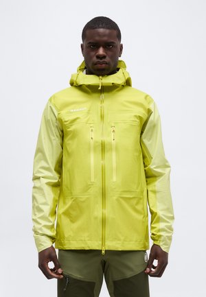 Mees, kes kannab erkkollast veekindlat kapuutsiga jopet, millel on kaks rinnaesist lukuga taskut, seisab ühevärvilise tausta ees.