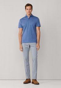 Polo bleu avec col, manches courtes et trois boutons, associé à un pantalon gris clair et des chaussures marron. Tissu à texture lisse.
