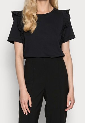 Femme portant un haut noir à manches courtes avec des détails à volants sur les épaules et un pantalon noir taille haute devant un fond uni.