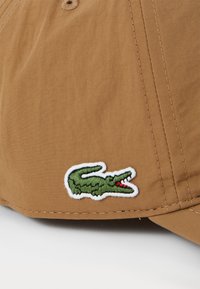 Casquette en coton marron avec un logo crocodile brodé vert et des accents rouges. La casquette présente un design structuré avec des coutures visibles.