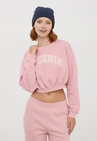 Felpa corta rosa con "PHOENIX" stampato in bianco. Abbinata a pantaloni della tuta coordinati e a un berretto di maglia blu scuro. Tessuto morbido.