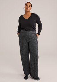 Mujer vestida con una camiseta negra de manga larga y escote en V, pantalones anchos grises texturizados, zapatillas negras y pendientes de aro en forma de corazón, de pie con las manos en los bolsillos.