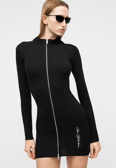 Γυναίκα που φοράει μαύρο μίνι φόρεμα με μακριά μανίκια, κουκούλα, φερμουάρ μπροστά και λευκό κείμενο κοντά στο τελείωμα, σε συνδυασμό με μαύρα γυαλιά ηλίου.