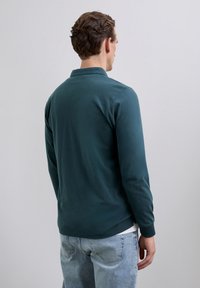 Hombre con el cabello corto y rizado, vistiendo una camiseta de manga larga color teal y jeans de un azul claro, de espaldas frente a un fondo gris liso.