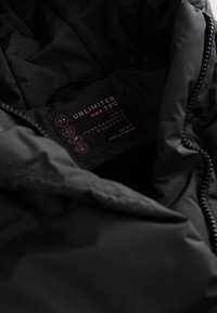 Gros plan de l'intérieur d'une veste noire montrant une étiquette avec "UNLIMITED TEC", des pictogrammes d'entretien et le texte "High Performance Garments".