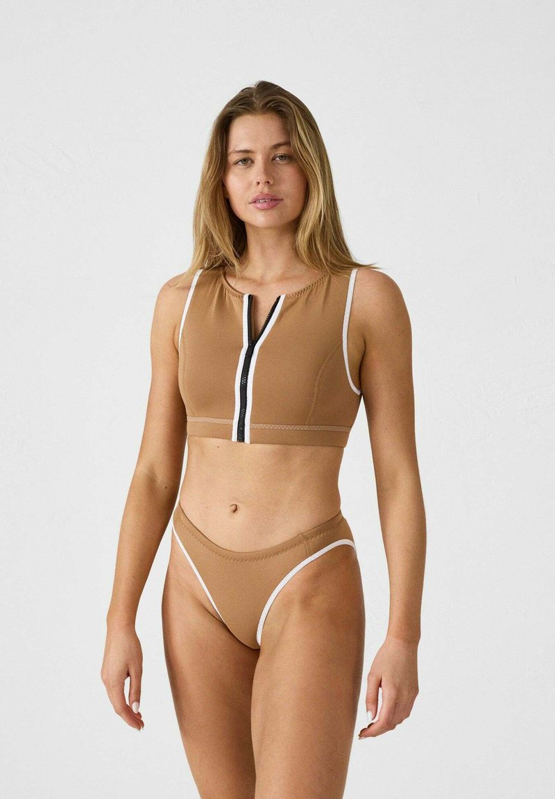 Top corto marrone con chiusura lampo davanti e dettagli bianchi, abbinato a uno slip bikini a vita alta coordinato, texture liscia, design minimal.