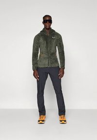 Olijfgroene fleece jas met ritssluiting, oranje accenten, gecombineerd met donkere broek en gele schoenen. Model staat tegen een effen achtergrond.