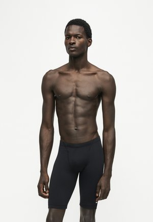 Uomo atletico con pelle scura che indossa pantaloncini neri aderenti, in piedi davanti a uno sfondo chiaro semplice, rivolto in avanti con espressione neutra.