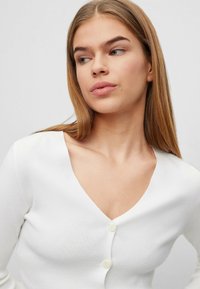 Weißer Cardigan aus geripptem Stoff, mit tiefem V-Ausschnitt, drei Knopfverschluss und taillierter Passform. Glatte Textur und klassische Silhouette.