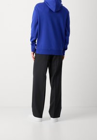 Blauwe hoodie met een trekkoordkap, geweven stof, gecombineerd met zwarte broek met wijde pijpen en witte sneakers. Gezien vanuit de rug.