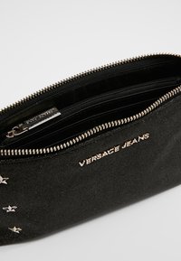 Pochette noire avec fermeture éclair, en tissu texturé, ornée d'accents en forme d'étoiles et d'un logo métallique "VERSACE JEANS".