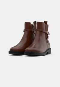 Rudos odiniai kulkšniniai batai su juodu guminiu padu, turintys diržą su auksiniu užsegimu ir elastinę įdėklą patogiam apsiaustimui.