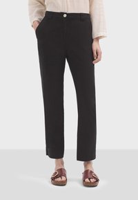 Schwarze, gerade geschnittene Hose mit Knopfverschluss, Seitentaschen und glatter Textur. Kombiniert mit braunen Sandalen mit einer runden Schnalle.
