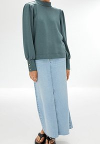 Jupe longue en jean bleu et haut vert à manches longues avec col montant, manches froncées et boutons dorés aux poignets.