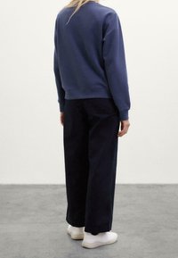 Persona vestida con un sudadera azul marino, pantalones anchos negros y zapatillas blancas, de pie sobre un suelo gris con un fondo blanco liso.