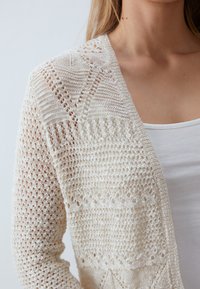 Detailní záběr ženy v krémovém, otevřeném pleteném kardiganu přes bílý tílko, ukazující jemné krajkové vzory na rukávu a přední části.