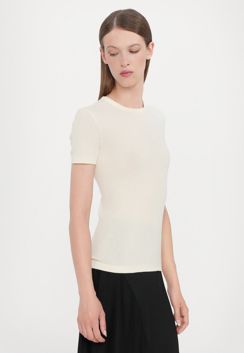Top de manga corta en color crema con un diseño ajustado, hecho de un material suave con un cuello redondo y una textura sutil. Combinado con una falda negra.