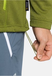 Zelená texturovaná bunda se zipem a nastavitelnou šňůrkou, v kombinaci se světle šedými kalhotami s bílými postranními zipy.