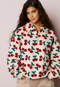 Pulover iz flisa s četrtinkim zadrgo, z kremno podlago in živahnimi rdečimi ter zelenkasto modrimi motivi češenj povsod. Mehka tekstura in sproščen kroj.