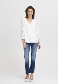 Weiße Bluse mit V-Ausschnitt und strukturiertem Blattmuster, kombiniert mit blauen Jeans. Das Modell steht vor einem neutralen Hintergrund und trägt cremefarbene Ballerinas.