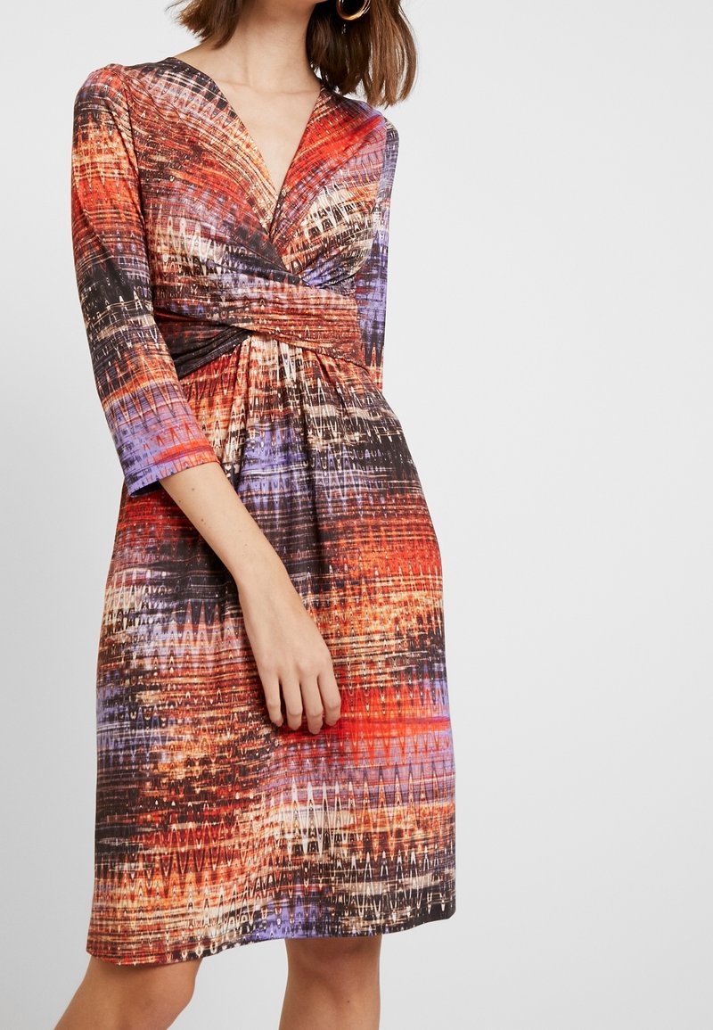 Femme portant une robe longueur genou avec manches trois-quarts présentant un motif abstrait multicolore en rouge, violet, noir et beige.