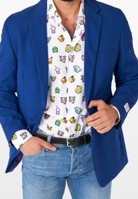 Blazer blu su una camicia bianca con pattern di personaggi dei cartoni animati colorati, abbinato a jeans blu chiaro e una cintura nera texturizzata.