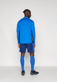 Ανδρική αθλητική ενδυμασία με μπλέ μπλούζα μακρυμάνικη με μαύρες πλευρικές πάνελ, μπλέ σορτσάκι, μπλέ κάλτσες μέχρι το γόνατο και μαύρα ποδοσφαιρικά παπούτσια.