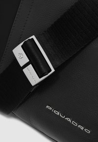 Schwarze Leder Tasche mit strukturiertem Finish, ausgestattet mit einer silbernen Schnalle an einem schwarzen Riemen und dem "Piquadro"-Logo, das auf der Oberfläche geprägt ist.