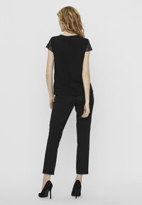 Mujer con cabello rubio ondulado que lleva una blusa negra con mangas de encaje, pantalones negros ajustados y tacones altos negros, de pie de espaldas sobre fondo blanco.