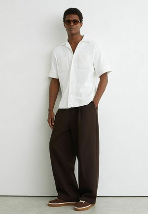 Homme portant une chemise blanche à manches courtes boutonnée, un pantalon large sombre, des baskets marron et des lunettes de soleil, debout contre un mur blanc.