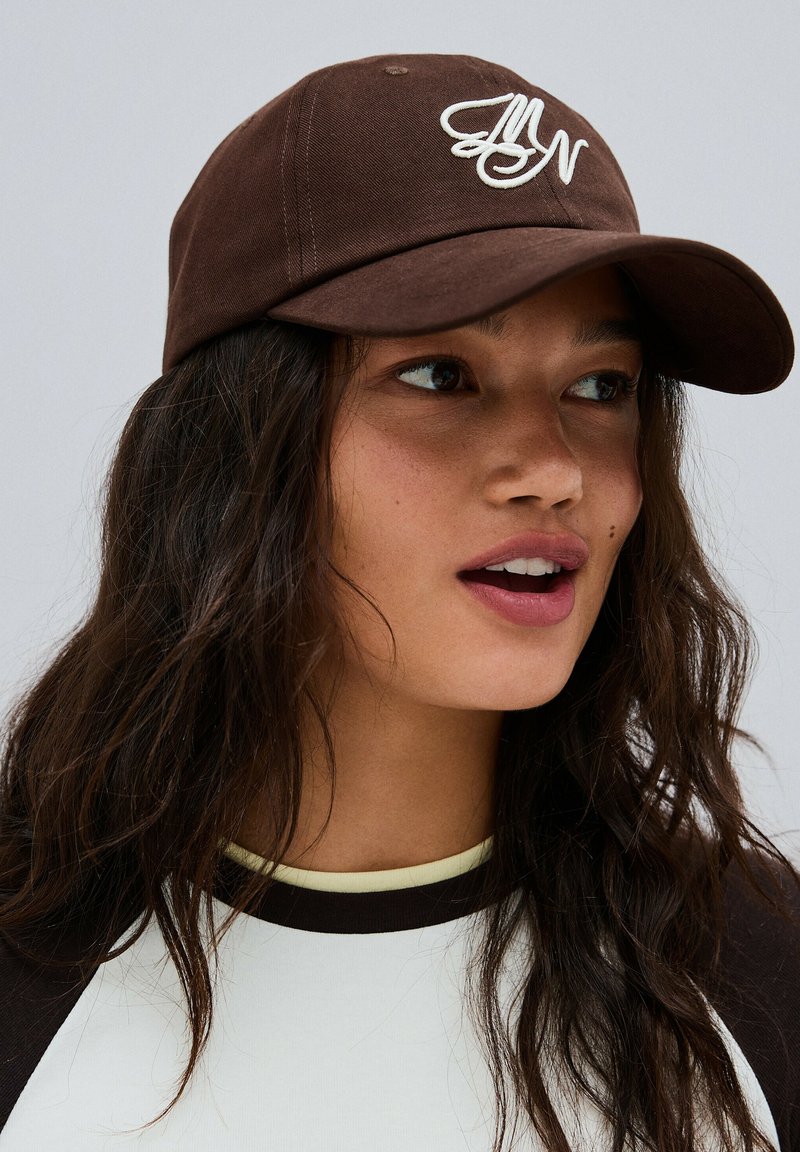 Jeune femme aux longs cheveux foncés portant une casquette marron avec un logo blanc et une chemise blanche à col noir, regardant sur le côté.