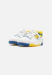 Zapatillas blancas con acentos en azul y amarillo, forro interior amarillo y detalle "550" en el costado, mostradas en ángulo sobre un fondo liso.