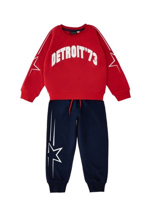 Felpa rossa a maniche lunghe con testo "DETROIT '73" e stelle bianche sulle maniche, abbinata a pantaloni blu navy con disegni di stelle bianche.