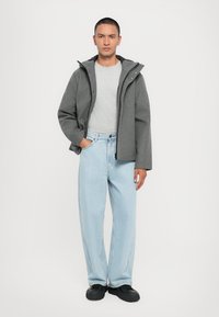 Palton gri din lână cu glugă, tricou gri, blugi albaștri deschis și adidași negri. Paltonul are un fermoar frontal și nasturi cu știft pentru închidere.