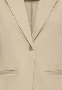 Blazer beige avec une fermeture à bouton unique, des revers crantés et deux poches horizontales à l'avant.