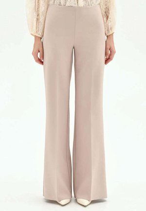 Pantaloni beige a gamba larga abbinati a scarpe bianche con punta a punta e un top lungo a maniche lunghe in pizzo color crema, modello in piedi con postura eretta.