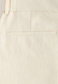 Gros plan d'un tissu texturé couleur crème avec des motifs floraux subtils et une poche passepoilée étroite.