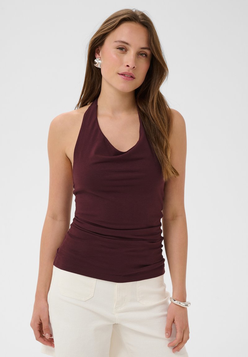 Femme aux longs cheveux bruns porte un top dos nu violet foncé, un pantalon écru, des boucles d’oreilles en argent et un bracelet en argent, debout sur fond blanc.
