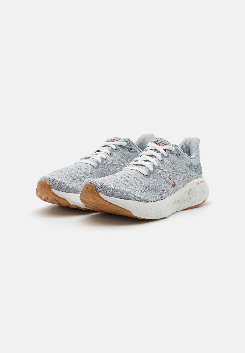 Een paar lichtgrijze hardloopschoenen met witte veters, witte tussenzolen en bruine gestructureerde buitenzolen schuin naar rechts op een witte achtergrond.