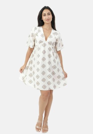 Kobieta w białej sukience o długości do kolan z beżowymi geometrycznymi wzorami, krótkimi rękawami i dekoltem w serek, ubrana w przezroczyste sandały na obcasie, stoi na tle jednolitego tła.
