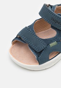 Blue suede sandal featuring an open-toe design, Velcro straps, and a textured beige insole. Accented with green stitching details.