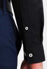 Camicia nera a maniche lunghe con polsini abbottonati, abbinata a pantaloni blu navy. La camicia presenta bottoni bianchi per la chiusura.