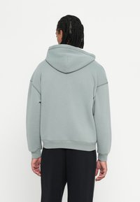 Person with braided hair wears light gray hooded sweatshirt and black pants, facing away against plain white background.